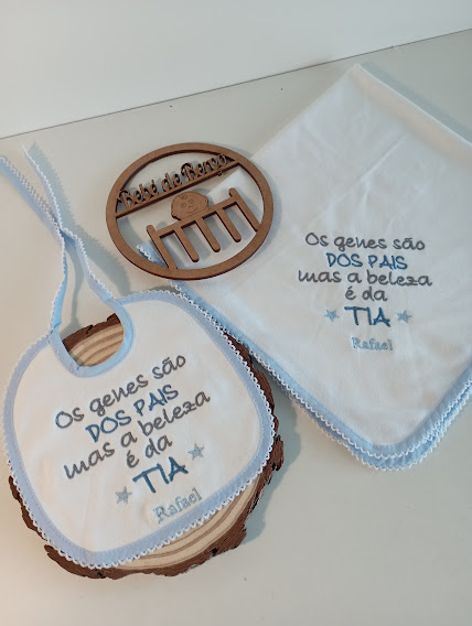 Babete e toalha bordados com texto em azul sobre superfície branca e base de madeira