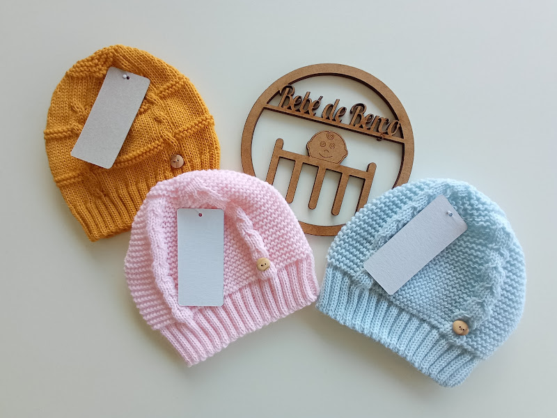 Três gorros de malha para bebé em mostarda, rosa e azul claro com etiquetas, botões de madeira e placa de madeira 'Pé de Benta'