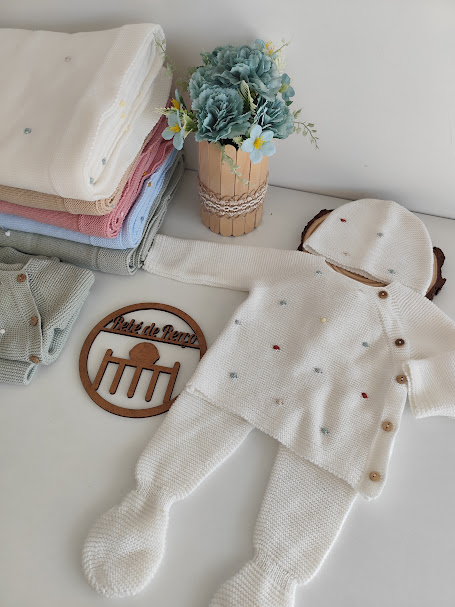 Conjunto de roupa de bebé de malha branca com botões e capuz, outras roupas coloridas, vaso de flores e placa de madeira.