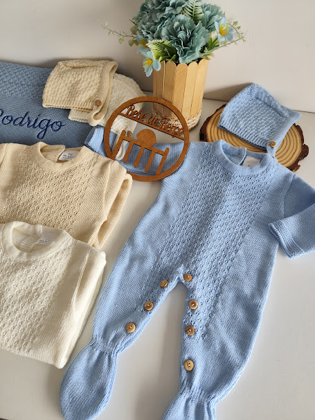 Conjunto de roupa de bebé em malha azul, bege e branco com botões de madeira, mantas bordadas com o nome Rodrigo e um vaso com flores
