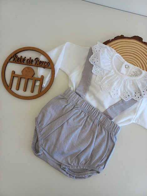 Conjunto de bebé branco com gola de renda e calções cinzentos com alças.