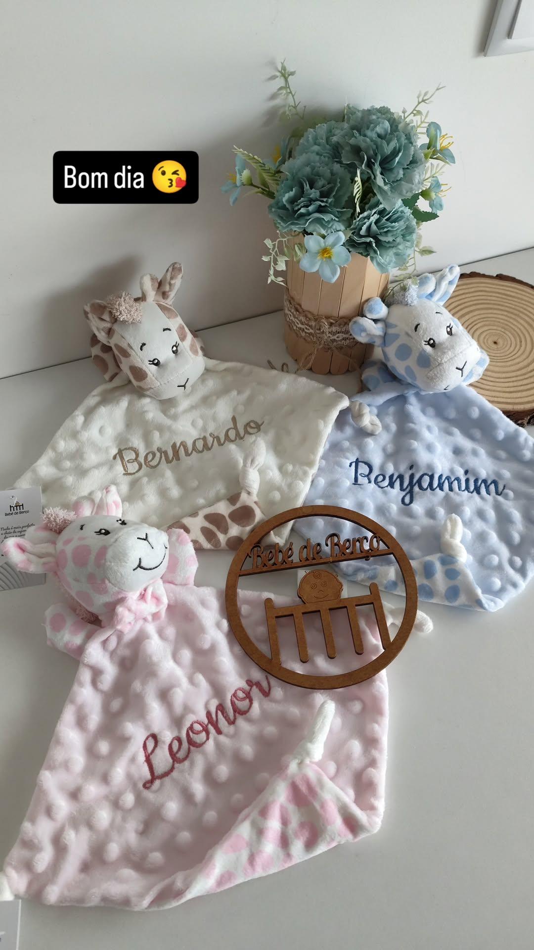 Três mantas de bebé com nomes bordados, cabeça de girafa como parte do design, flor azul artificial em vaso e decoração de madeira gravada.