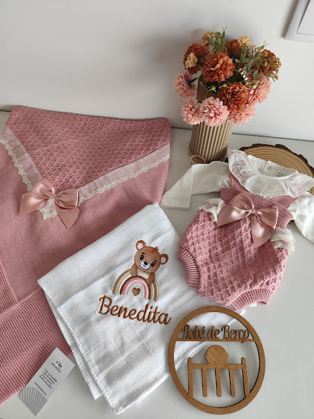 Conjunto de roupa de bebé rosa com laços, manta e toalha bordada, e placa decorativa em madeira com vaso de flores
