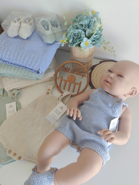 Boneco bebê com macacão azul claro de malha e sapatinhos, rodeado por macacões e sapatinhos de malha, manta azul, flores e placa decorativa em madeira