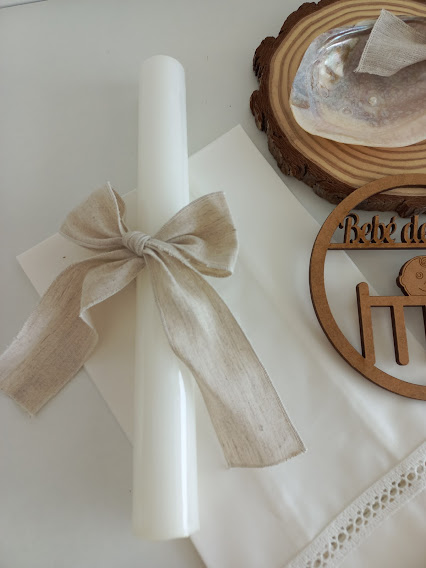 Vela branca com laço bege em tecido junto a suporte de madeira com texto recortado e concha prateada
