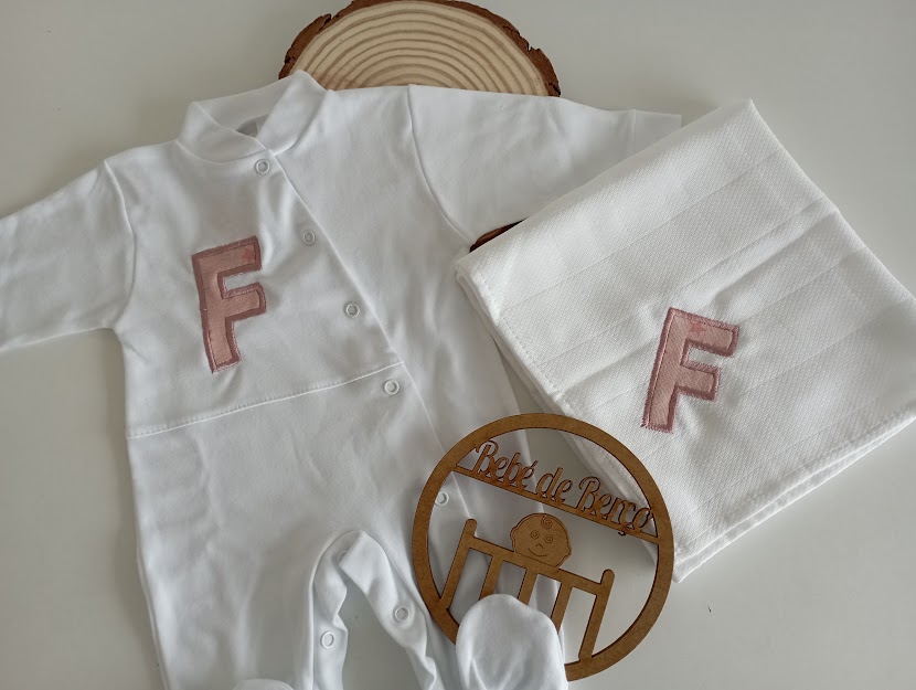 Conjunto branco de roupa de bebé com letra F rosa e manta em fundo branco com elementos decorativos em madeira