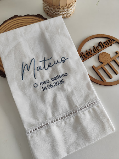 Toalha branca bordada para batismo com nome e data, sobre superfície branca com objetos decorativos de madeira