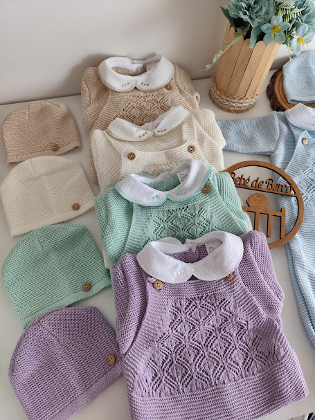 Conjunto de roupa de bebé em diferentes cores com detalhes bordados e botões, acompanhado de gorros e decoração de flores