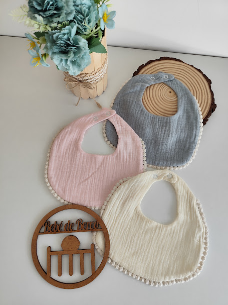 Três babetes de algodão cor cinza, rosa e creme com pompons, arranjo floral e ornamento de madeira