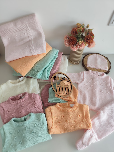 Conjuntos de roupa de bebé em malha colorida pastel com padrão texturizado, mantas, gorros e flores num vaso sobre mesa branca