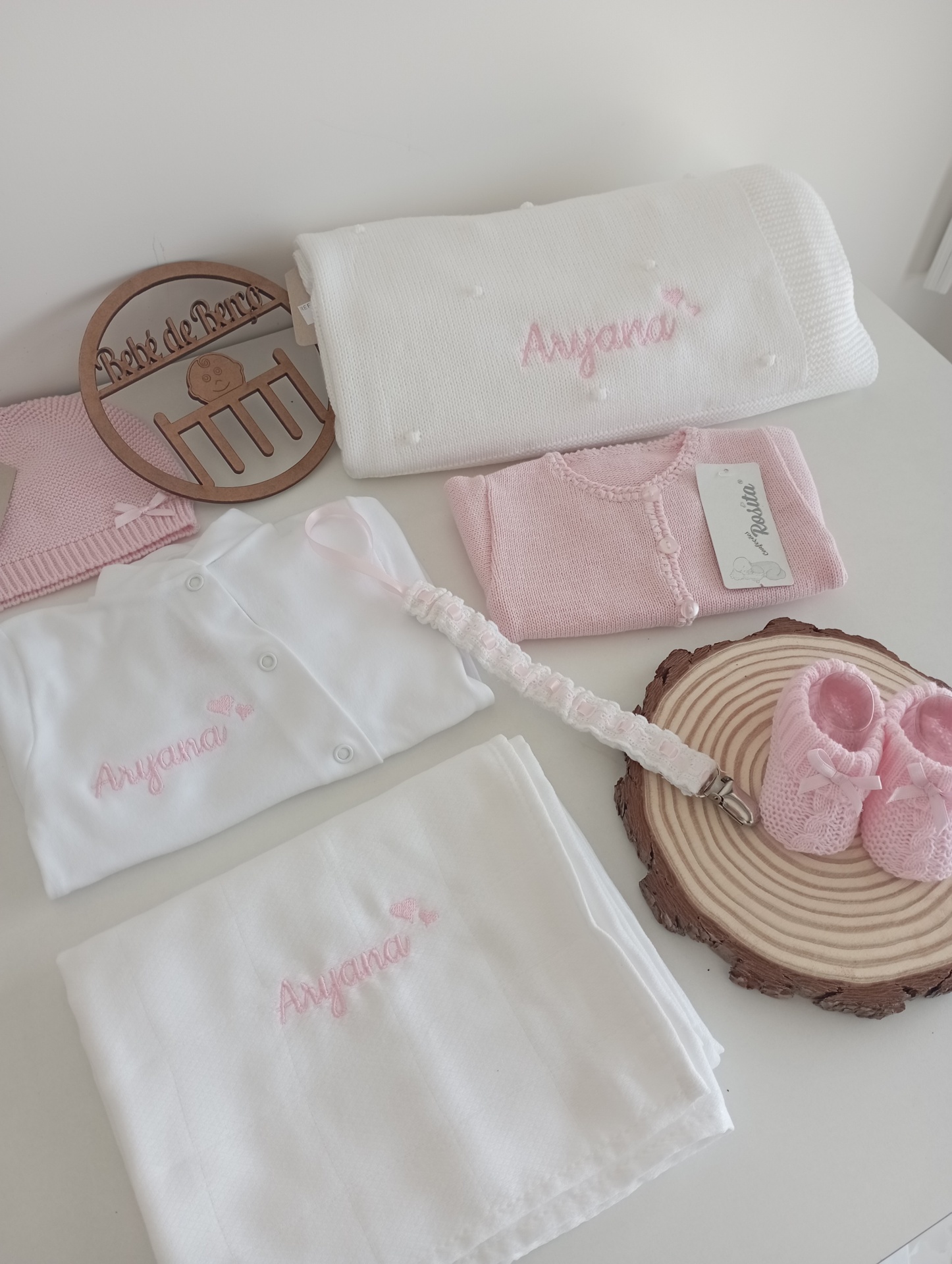 Conjunto de roupa e acessórios de bebé com bordados 'Anyana' em branco e rosa.