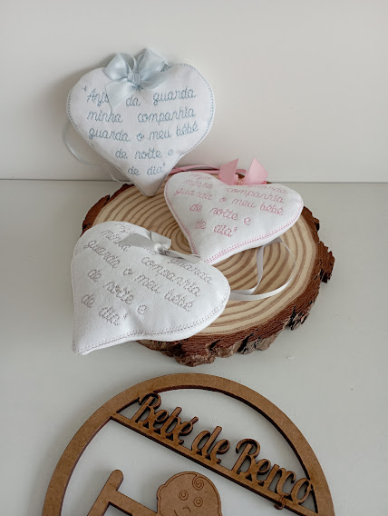 Três almofadas de tecido branco em forma de coração com bordados coloridos e texto, sobre base de madeira redonda, e ornamento de madeira com texto.