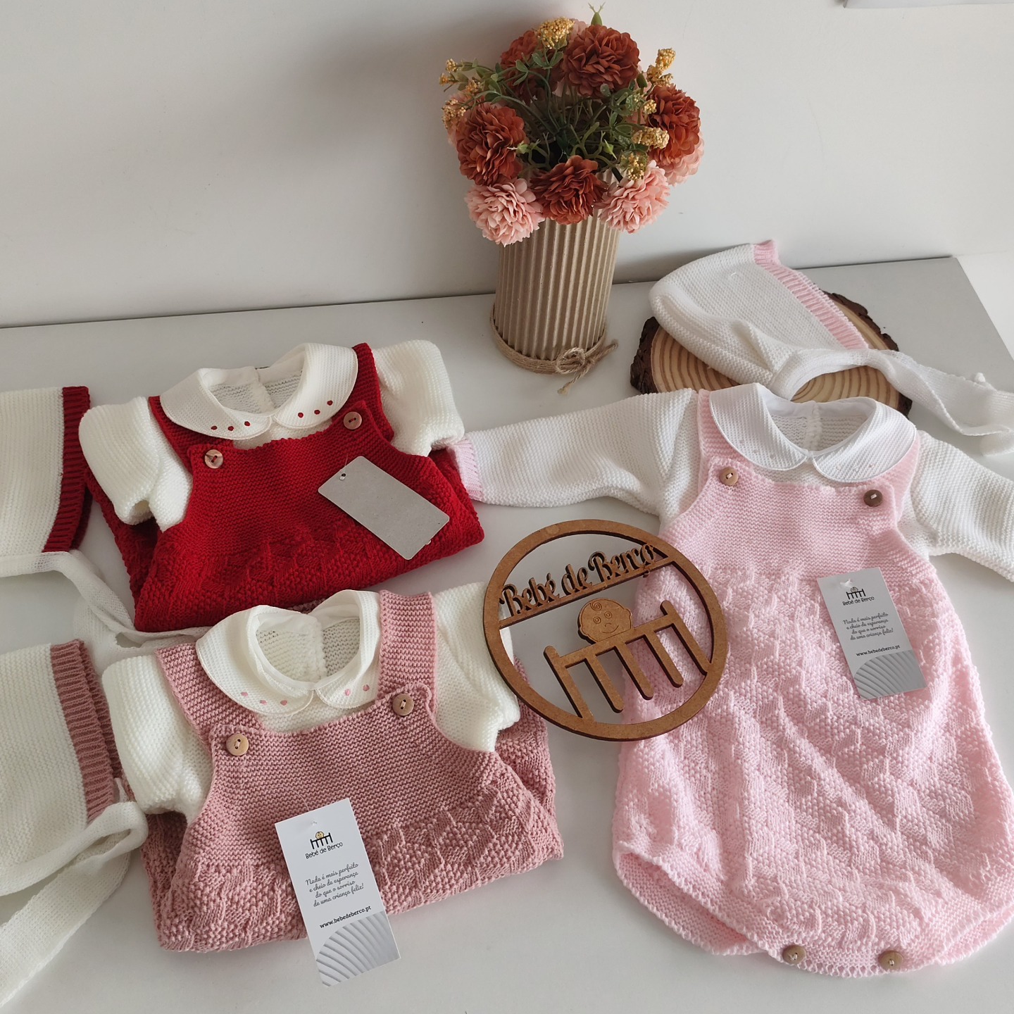 Conjuntos de roupa de bebé em malha vermelha, rosa claro e branca, sobre mesa branca com vaso de flores secas