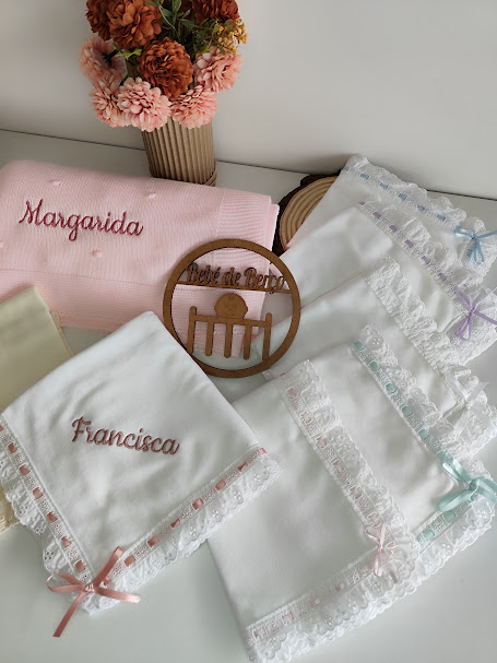 Toalhas de bebé bordadas com rendas e fitas em cores suaves ao lado de vaso com flores secas