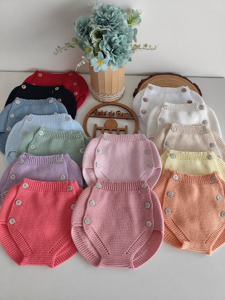 Conjunto de roupa de bebé em malha com botões laterais em várias cores sobre mesa branca com vaso de flores e placa de madeira