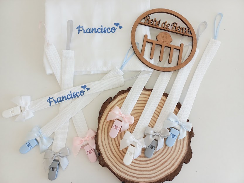 Conjunto de chupetas com fitas de tecido branco e laços coloridos, nome Francisco bordado em pano branco e placa redonda de madeira.