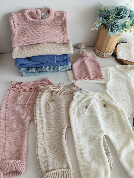 Conjunto de roupa de bebé em malha colorida com botões de madeira e gorro rosa, fundo branco e vaso com flores azuis