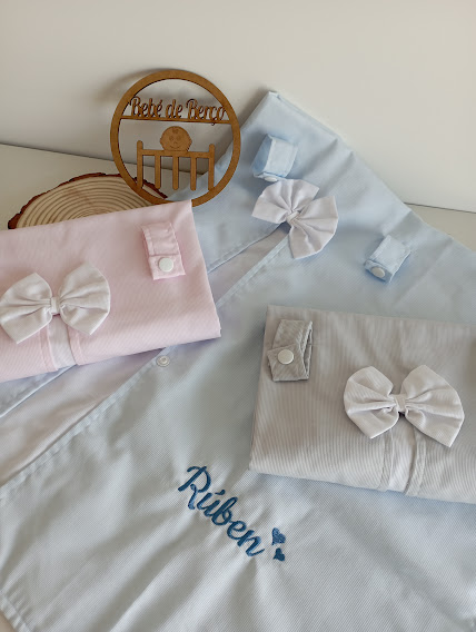 Roupas infantis em azul, rosa e cinza com laços brancos e nome bordado Rúben sobre fundo claro e disco de madeira Bebé de Peso