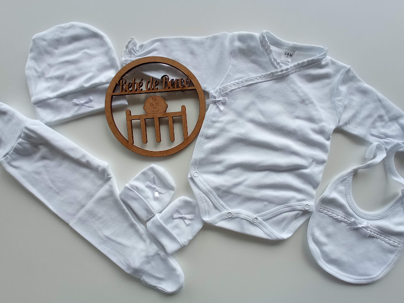 Conjunto branco de roupa de bebé com body, calças, babete, touca e peúgas ao lado de enfeite de madeira redondo.