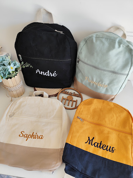 Quatro mochilas coloridas com nomes bordados e um vaso de flores em fundo branco