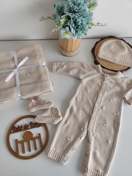 Conjunto de roupa de bebé bege com bolinhas brancas e manta, com vaso de flores azuis e placa de madeira com texto