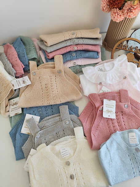 Conjuntos de roupa de bebé em malha de cores suaves organizados numa mesa branca com jarra de flores cor-de-rosa ao fundo