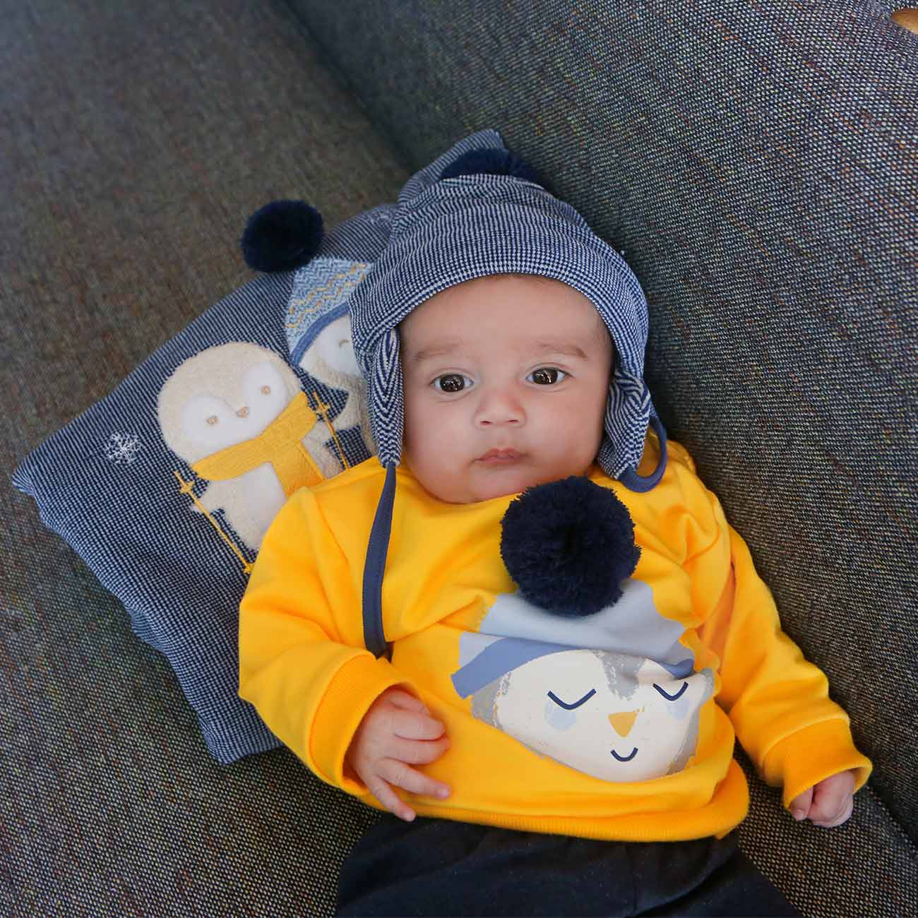 bebé com camisola amarela e gorro azul deitado num sofá com almofada de pinguins