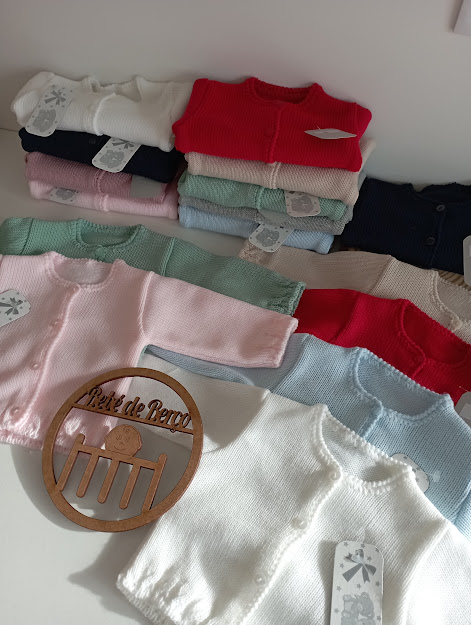 Cardigans de bebé de várias cores empilhados com letreiro Petit de Bébé
