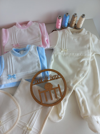 Roupa de bebé branca, azul, rosa e bege com renda, rolos de linha colorida e recorte de madeira com texto