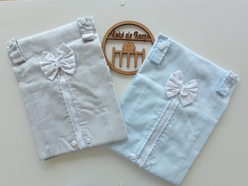 Dois panos bordados para bebé, um branco e um azul claro, com laços, sobre superfície branca e ornamento de madeira