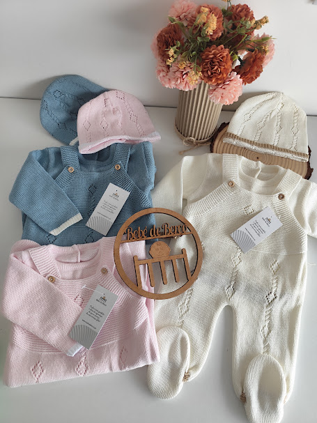 Conjuntos de roupa de bebé de malha azul, rosa e branca com gorros e ornamento de madeira, junto a jarro com flores artificiais.