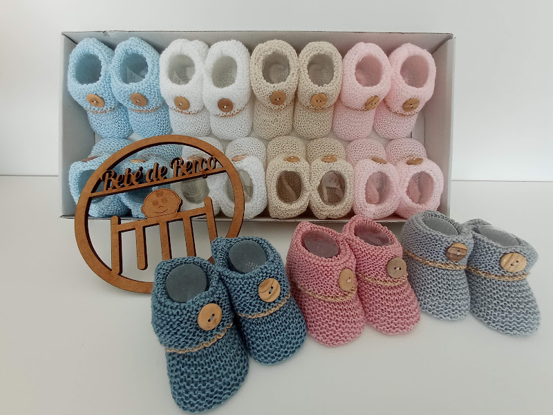 Vários pares de sapatinhos de malha para bebés com botão de madeira, organizados em caixa branca, com placa decorativa em madeira com texto Pézinhos de Lã.