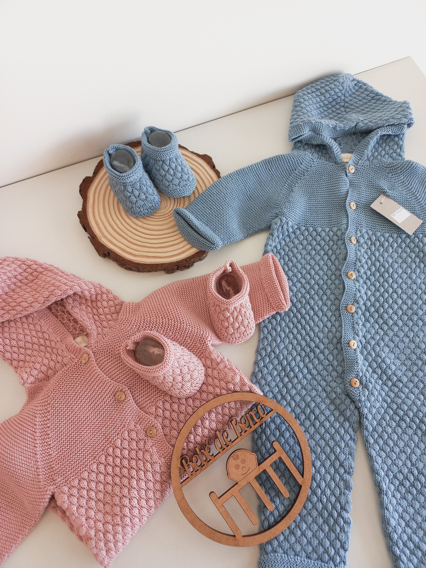 Dois macacões de bebé em tricô azul e rosa com botinhas combinando, dispostos sobre superfície clara e fatia de madeira decorativa