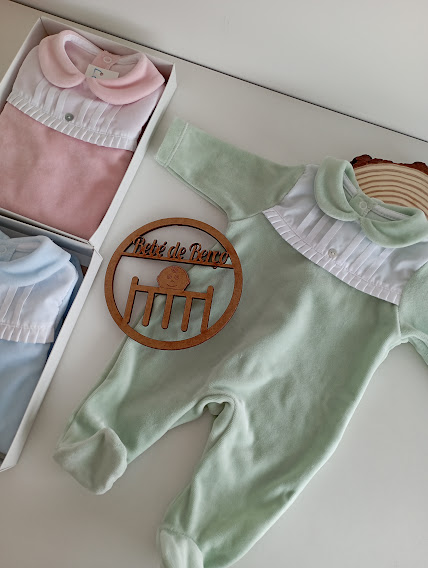 Fatos de bebé em veludo verde, rosa e azul claro com colarinhos e pregas brancas, placa de madeira com texto na superfície branca.