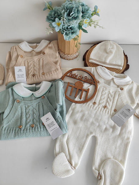 Roupas de bebé em malha bege, verde e branca com padrões trançados e boina branca ao lado de um vaso de flores azuis
