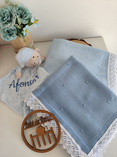 Manta azul com renda, pano de boca bordado Afonso, boneco de pelúcia e flores num vaso