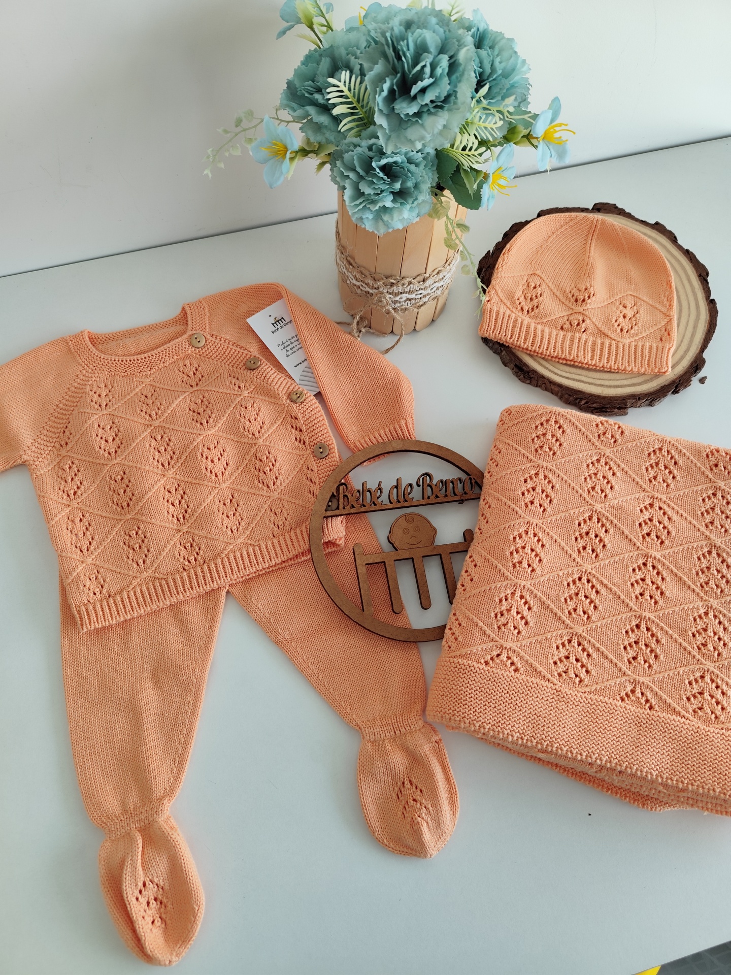 Conjunto de roupa de bebé cor pêssego em malha com casaco, calças, boina e manta com padrão floral em losangos, e vaso de flores azuis sobre superfície branca