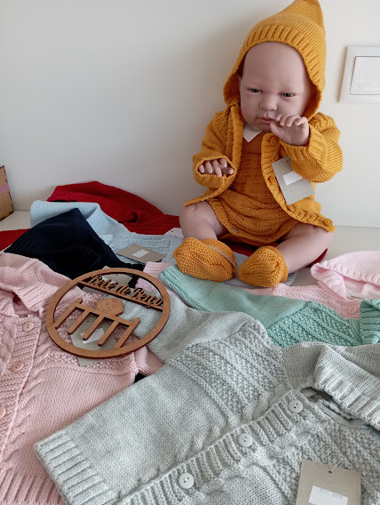 Boneca com roupa de malha mostarda com várias roupas de malha em cores pastel e vermelhas à sua volta