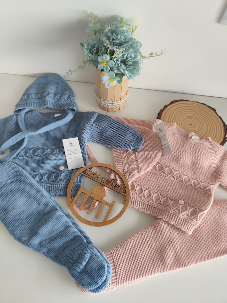 Conjuntos de roupa de bebé em malha azul e rosa com padrão de tranças, vaso com flores e enfeite de madeira