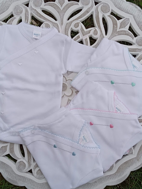 Cinco bodies de bebé brancos com bordados de corações e detalhes coloridos sobre fundo decorativo branco