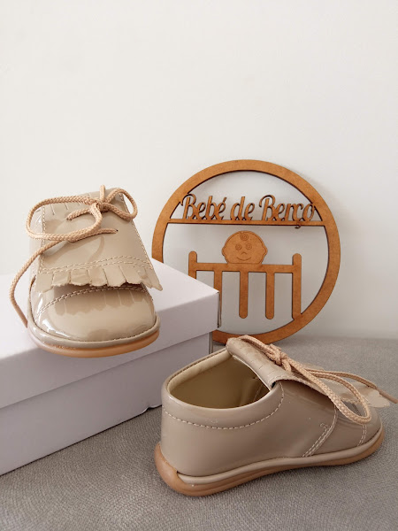 Sapatos de bebé em verniz bege com franjas e cordões sobre caixa branca, placa decorativa em madeira com texto 'Bebé de Berço' no fundo.