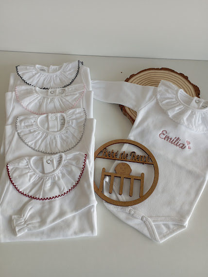 Bodies brancos de bebé com golas frisadas e detalhes de bordado em várias cores, incluindo um body com o nome Emília bordado em rosa.
