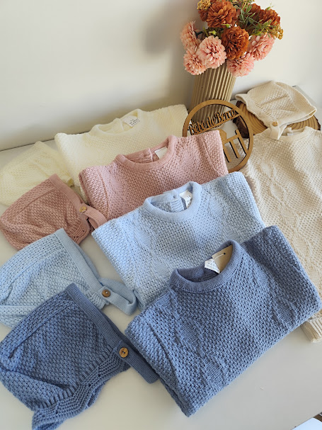 Conjunto de roupa de malha para bebé em tons creme, rosa e azul com flores secas num vaso