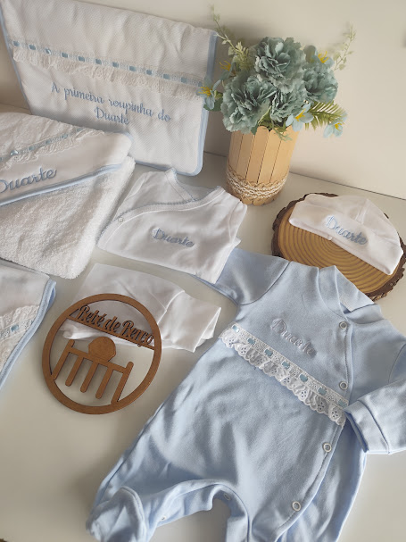 Conjunto de roupas de bebé branco e azul claro com bordados e detalhes em renda, junto de flores e objeto de madeira