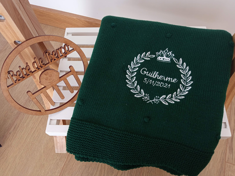 Cobertor verde escuro com bordado branco e cabide de madeira pendurado na cadeira branca