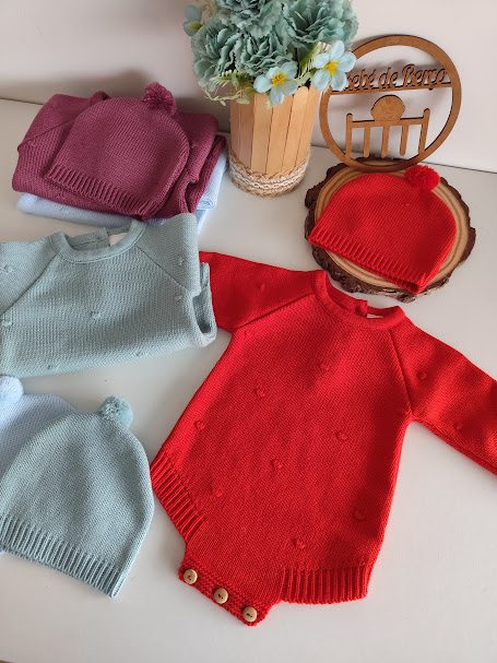 Body de malha vermelho e gorro com pompom, outros bodies e gorros em azul e vinho, vaso com flores e placa de madeira com texto