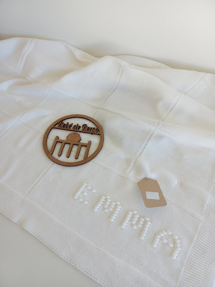 Manta branca com padrão quadriculado, palavra EMMA em relevo, etiqueta castanha e emblema redondo de madeira