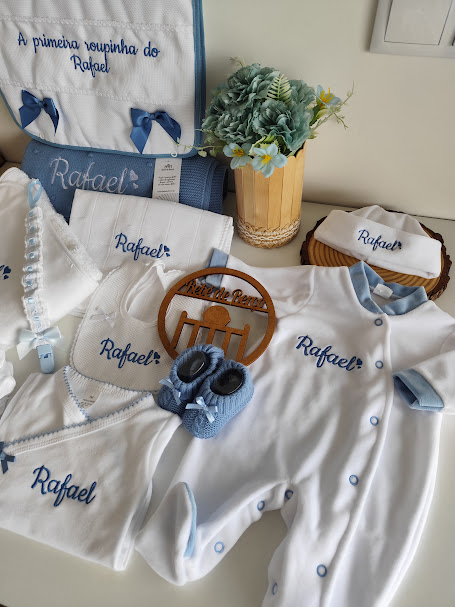 Conjunto de roupa de bebé branco com bordados azuis e nome Rafael, com acessórios e flores azuis ao fundo