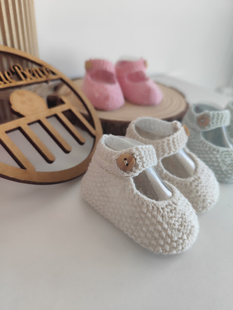 Sapatinhos de bebé em crochet creme, verde água e rosa com botões de madeira, sobre superfície branca e placa de madeira ao fundo