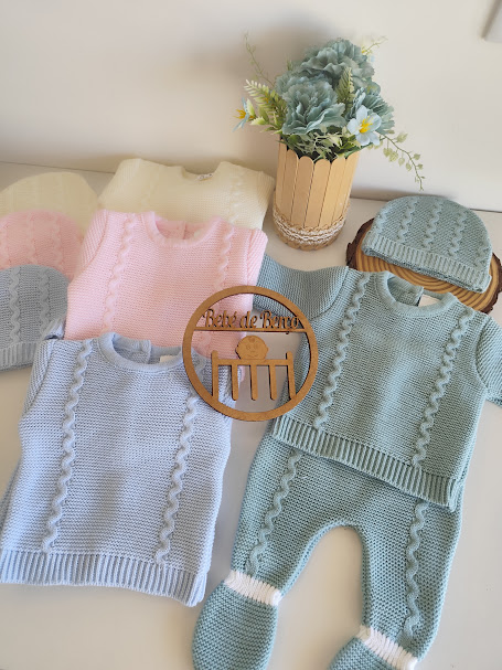 Conjunto de roupa de bebé em tricô azul, rosa e bege com padrão trançado e placa redonda de madeira com texto
